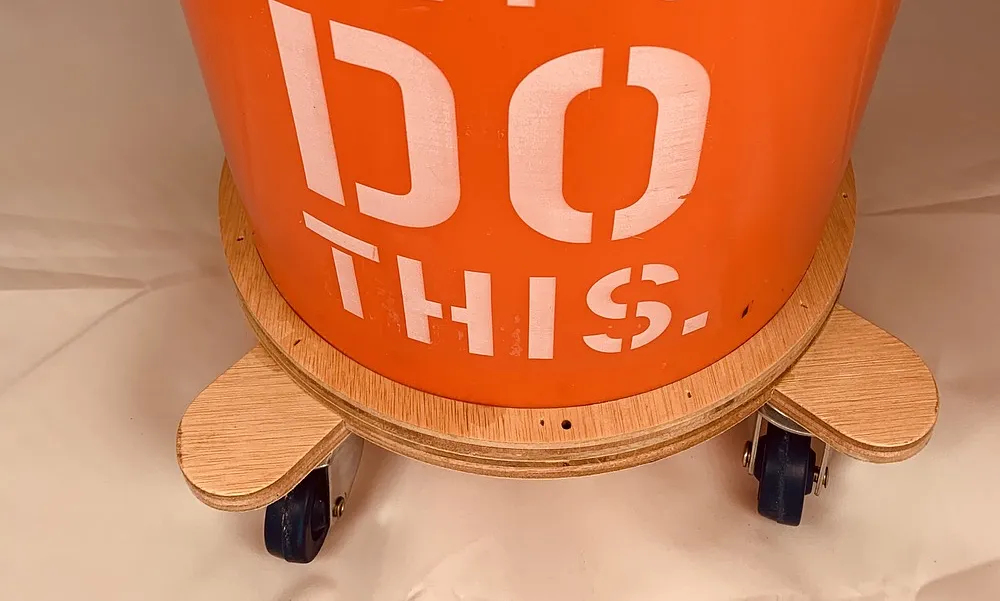 Orange five-gallon bucket sitting on a plywood dolly with caster wheels